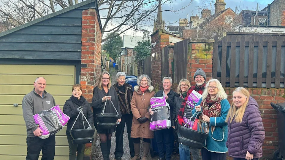 10 people in a line by a alley way they are holding plant pots and gardening soil.