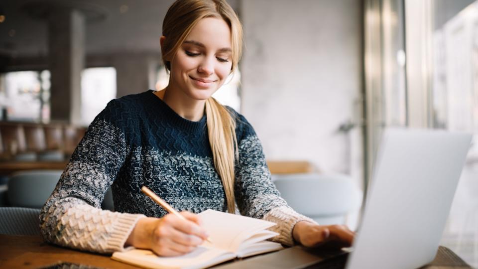 Young attractive university student using laptop computer, studying online at modern library. Cheerful caucasian woman writes notes, planning working process, sitting in loft cafe. Exam preparation