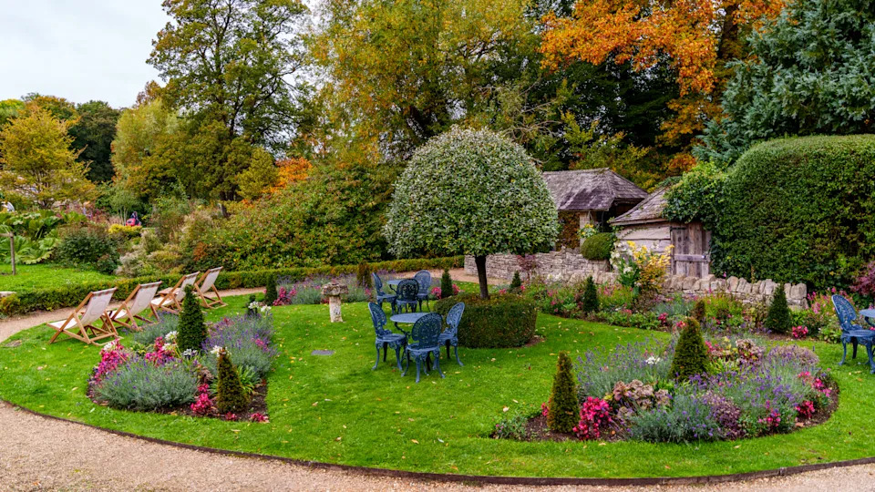 Lush formal garden featuring cast iron tables with chairs, deckchairs, colorful flowering plants, manicured lawn, and autumn foliage
