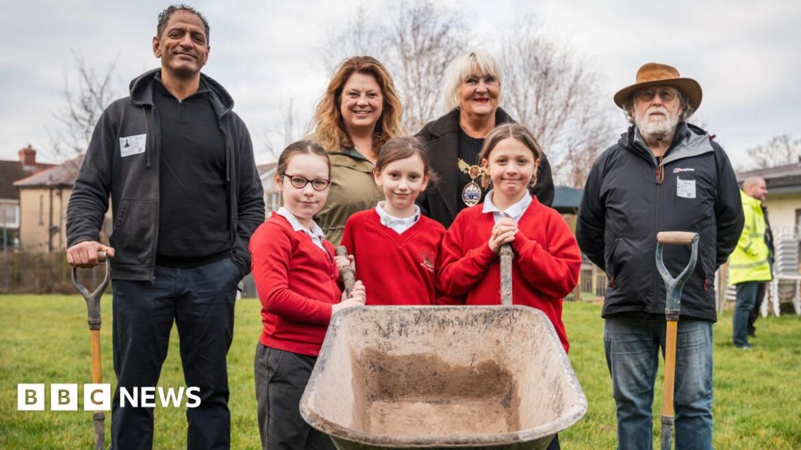 Two men and two women are standing next to three schoolchildren who are holding a wheelbarrow. The school and the school field can be seen behind them.