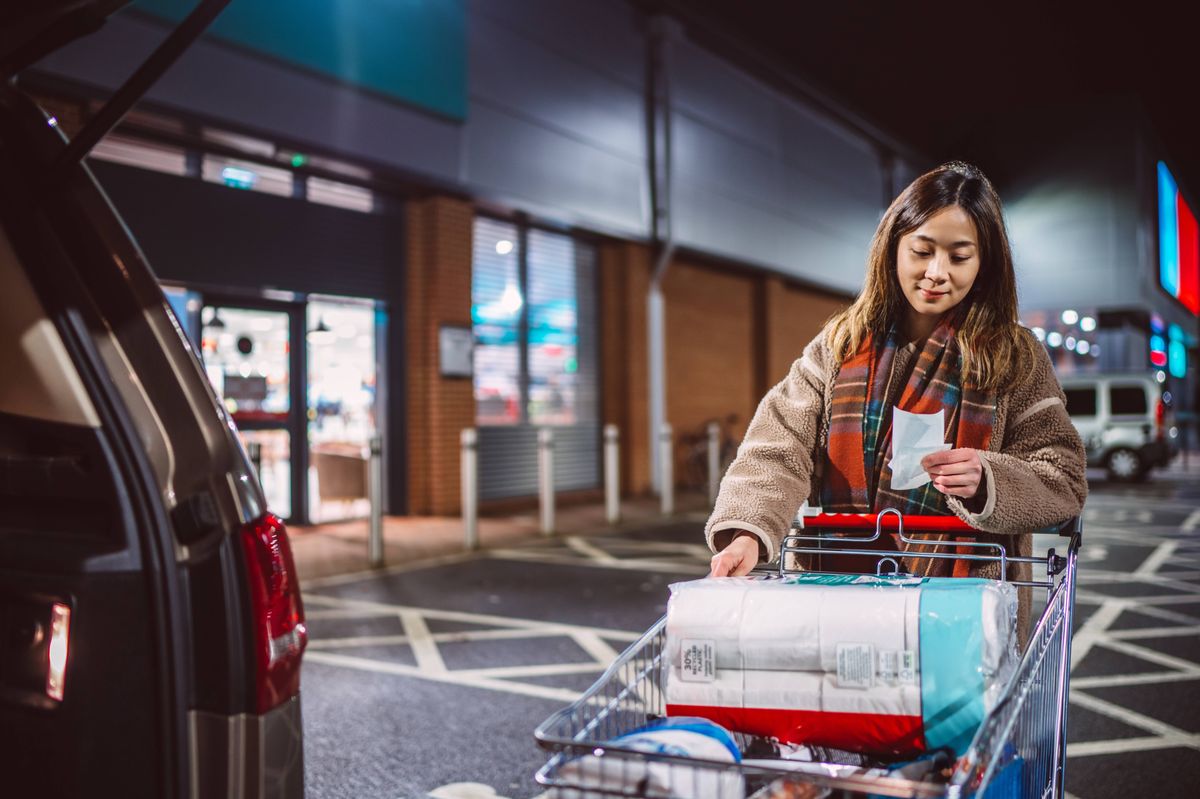 Woman checking her shopping receipt beside a supermarket parking lot at night, loading household essentials into her car.