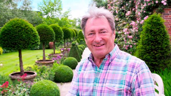 An older gentleman, wearing a plaid shirt, is standing in a well-maintained garden filled with neatly trimmed hedges and topiary