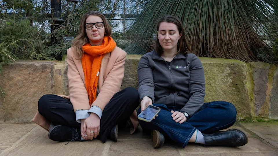 Two women, sitting on the ground with their legs crossed. The woman on the right is holding a phone in her hand and is wearing a grey top, blue jeans and has black boots on. She is looking down. The other woman has her eyes closed, is wearing glasses and has a pink jacket on with an orange scarf, black trousers and dark boots. They are both sat in front of plants. 