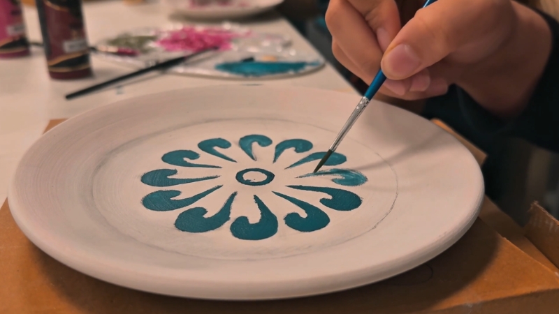 Hand painting a blue floral pattern on a ceramic plate with a small brush