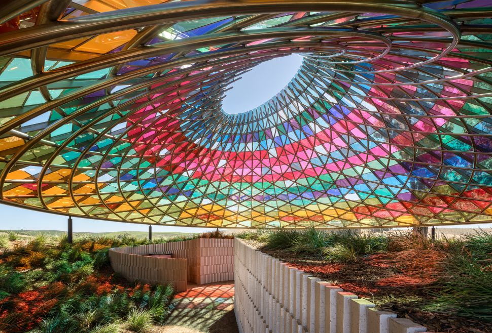 View from inside Olafur Eliasson’s Vertical Panorama Pavilion, showing a circular opening framed by a rainbow-colored glass canopy above landscaped paths.