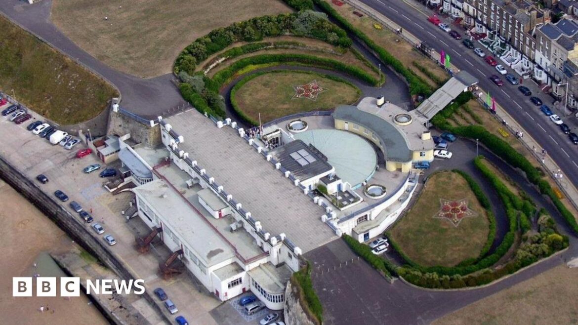 An aerial shot of a circular building surrounded by gardens and bordered by a road and a beach.