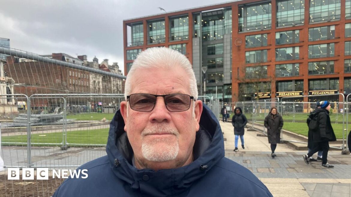 A man with short grey hair and a beard is wearing glasses and a dark blue coat while standing close to railings in Piccadilly Gardens. Around six people can be seen walking nearby.