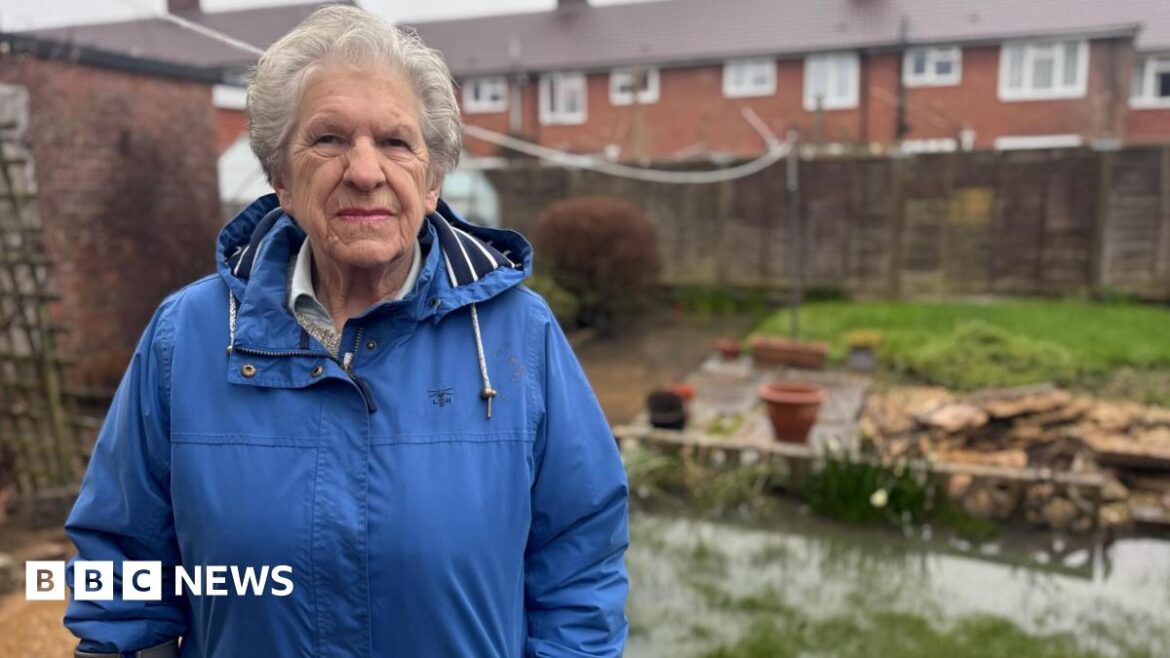 An elderly woman in a blue coat, stood to the left of her garden which is partially submerged underwater by the rain. She is stood in front of a brown fence and a row of houses.