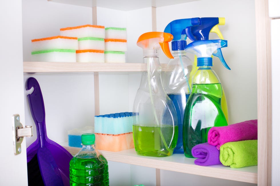 cleaning supplies and tools arranged on shelves in pantry including stacked sponges, spray bottles, rolled brightly colored cloths and a dust pan
