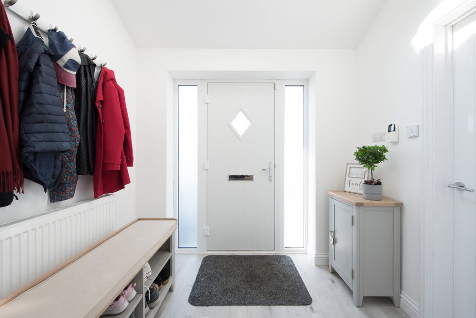 a general interior view of a hallway painted white, with front door, doormat, grey console cabinet, potted plant, coat rack with coats, shoe rack bench