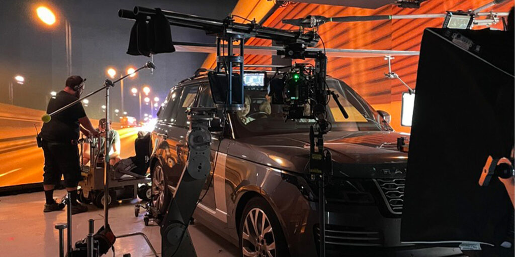 A film crew at Garden Studios operates cameras and lighting equipment around a parked SUV in a studio, with a blurred city lights background projected behind the vehicle.
