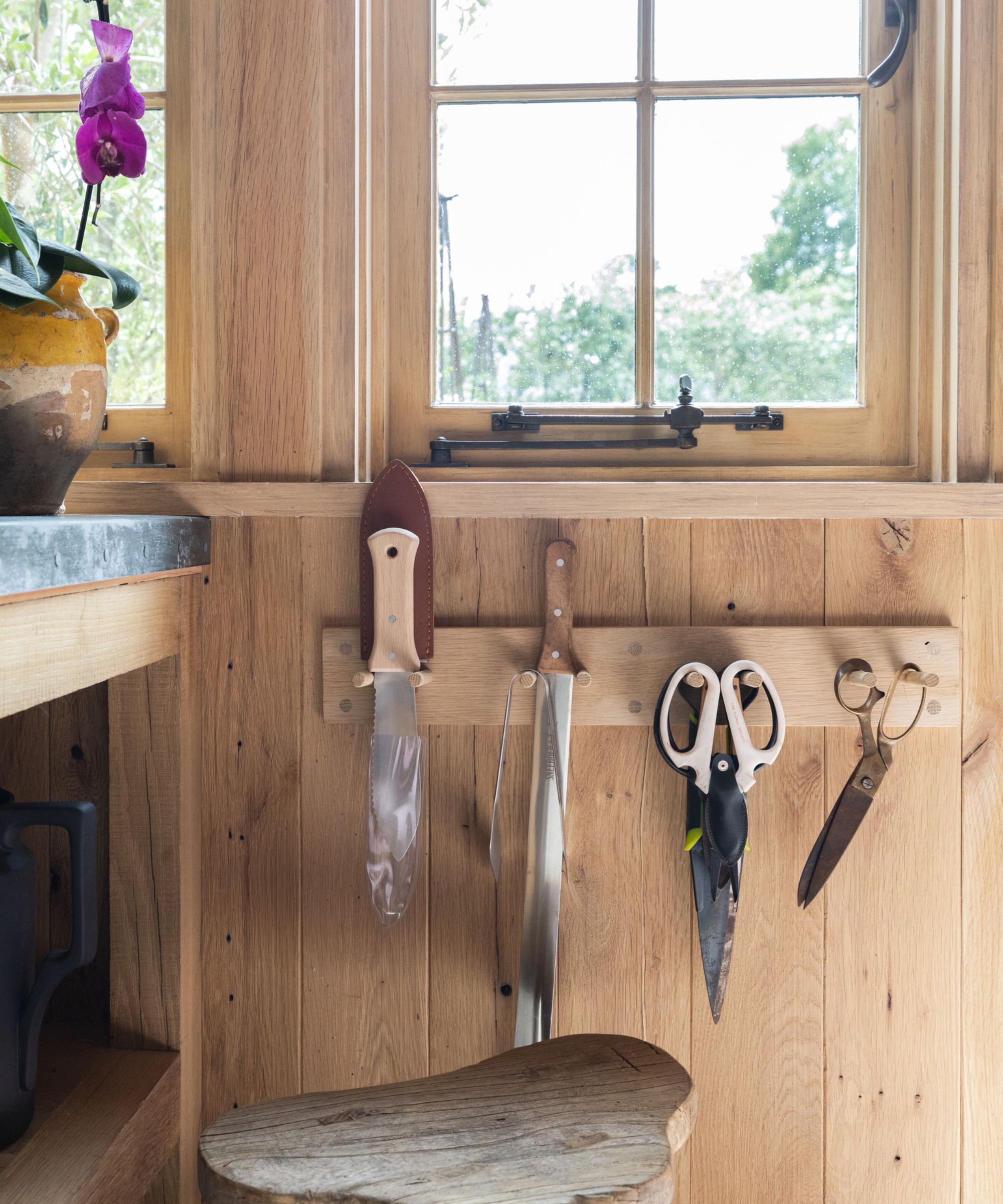 A close up of a small wooden peg rack on a wooden shed wall, with trowels, knives, and scissors hanging from it.