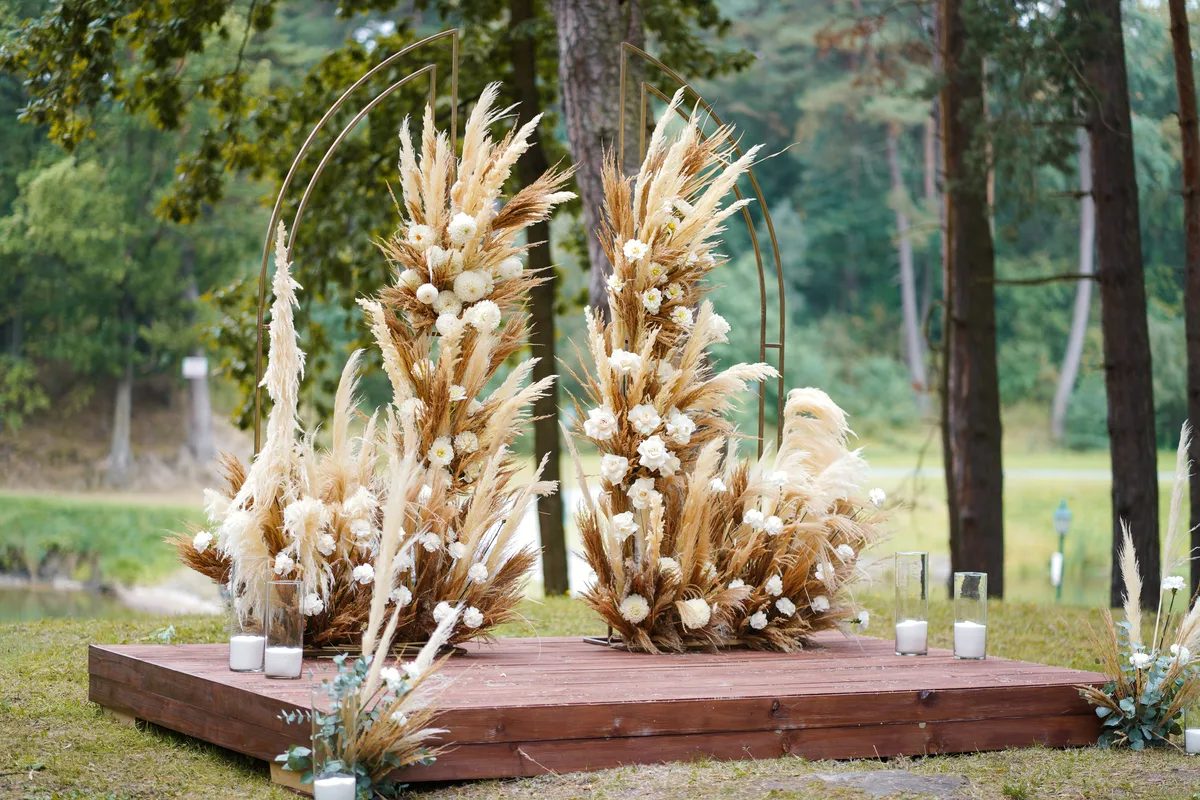 Wedding arch with pampas grass and white flowers on wooden stage in the forest, decorated with candles.