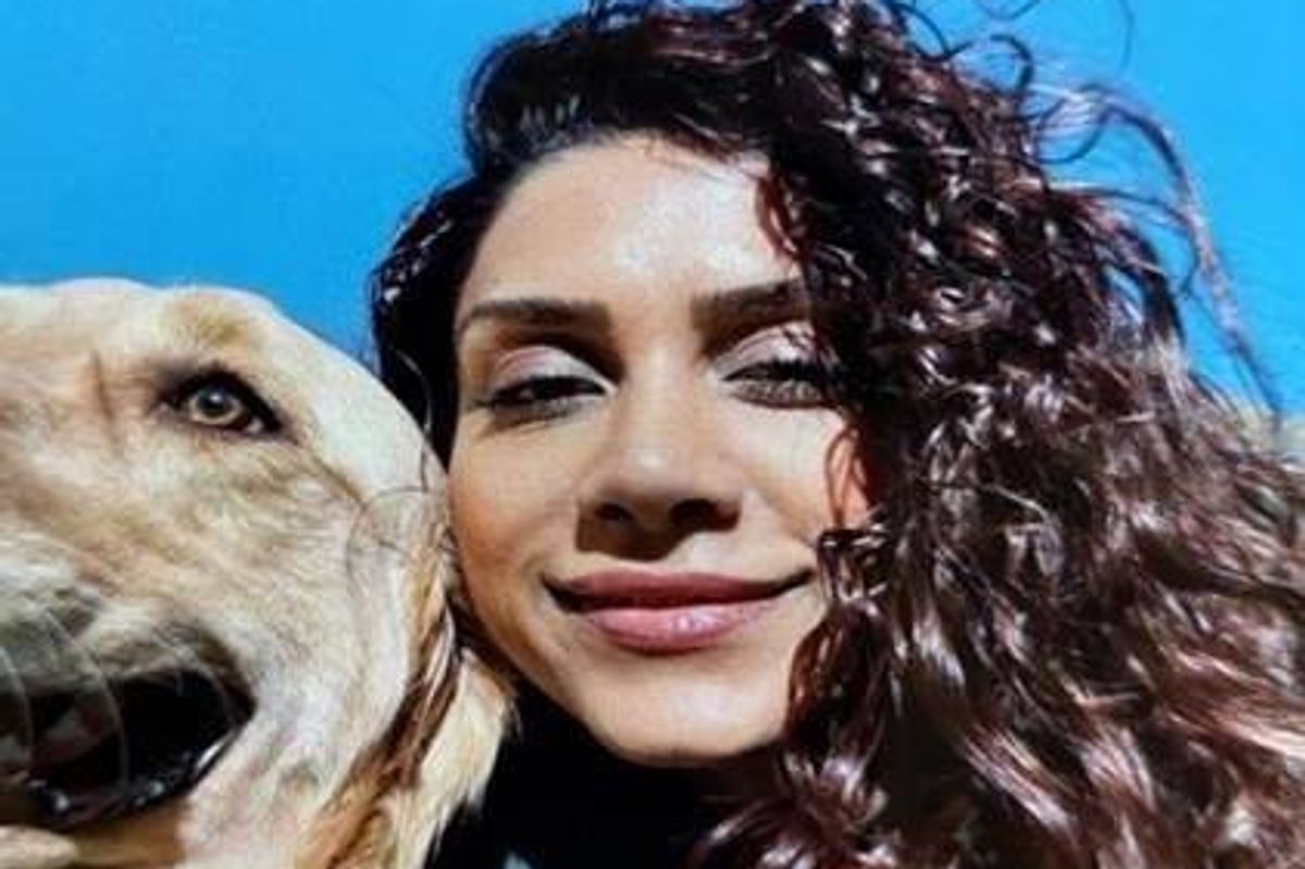 An individual with long curly hair is standing beside a golden retriever dog, posing for a photograph with a blue background.