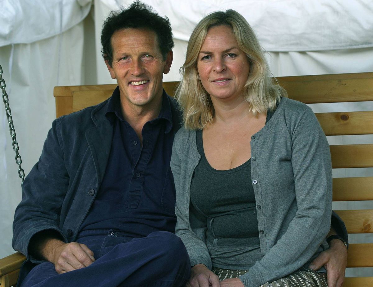 Two individuals, a man and a woman, are seated together on a wooden bench, both wearing casual attire and smiling, as if posing for a photograph against a backdrop of a white tent.
