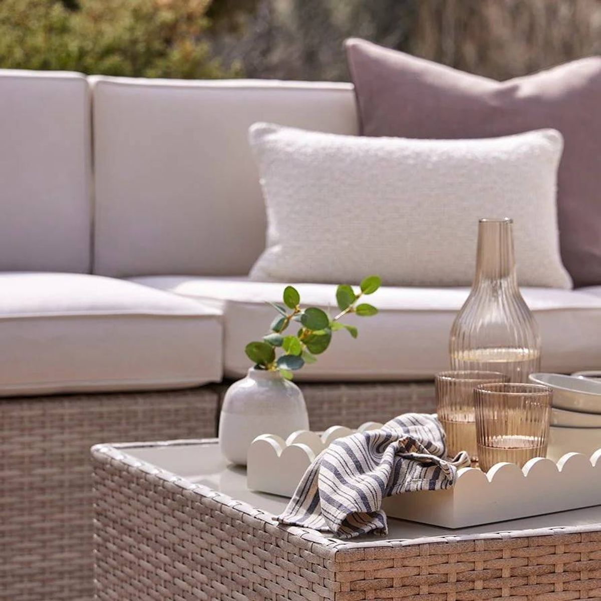 An elegant living room setting features a beige sectional sofa adorned with white cushions. A decorative tray rests on a wooden coffee table, holding a clear glass vase with a single green plant, a translucent bottle, and a striped cloth. The ambiance is complemented by the soft natural light filtering into the room.