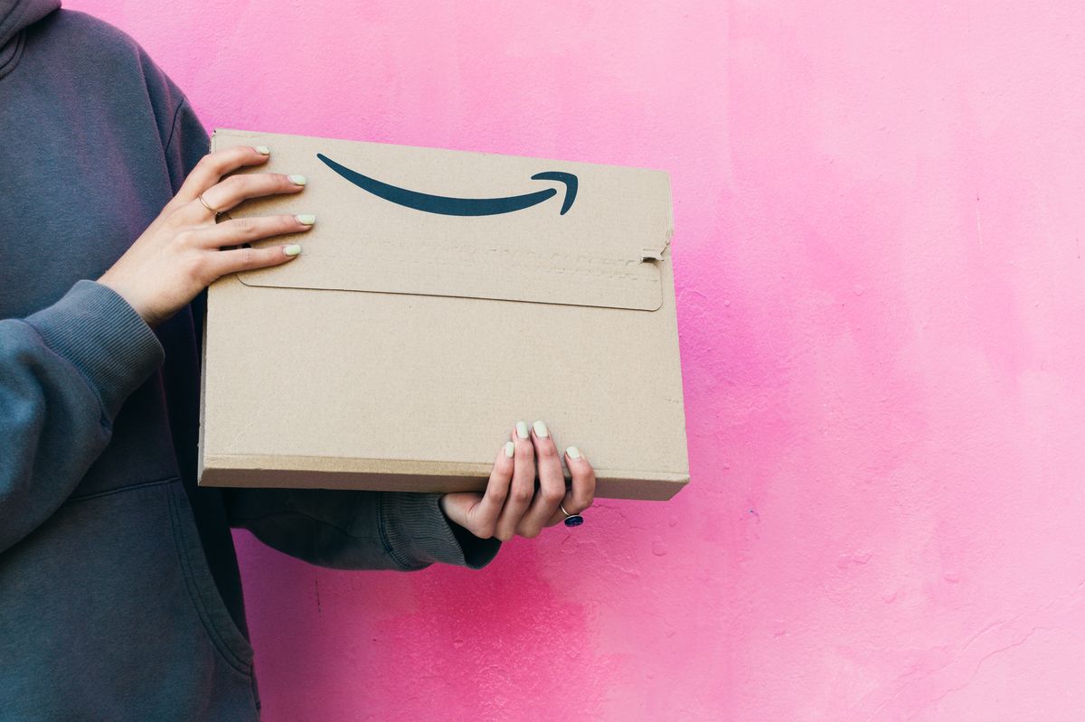 body of young woman holding an amazon packet on pink background. The package is made of cardboard and has the amazon symbol in blue. The image was made indoors when you received the package at home. It's about online shopping. The person cannot see his face and is unrecognizable.