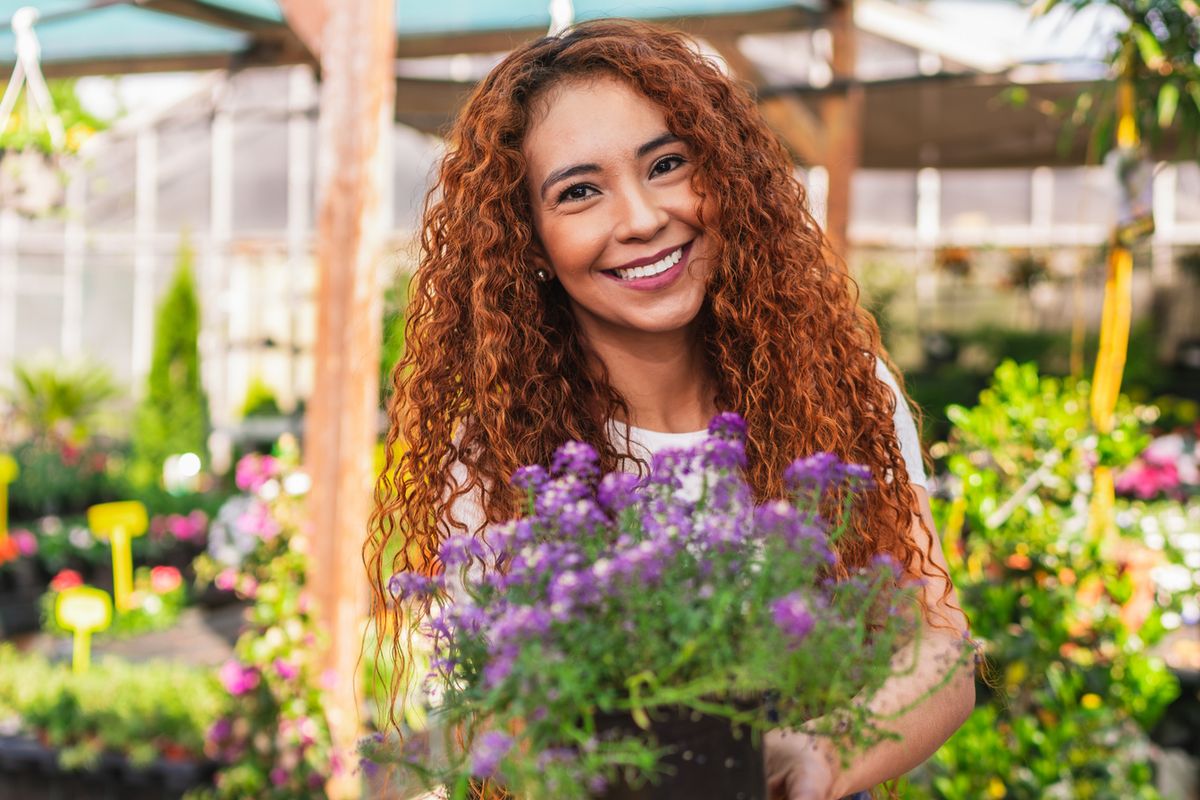Happy latin american woman working in a plant nursery, holding a flower pot and smiling at the camera