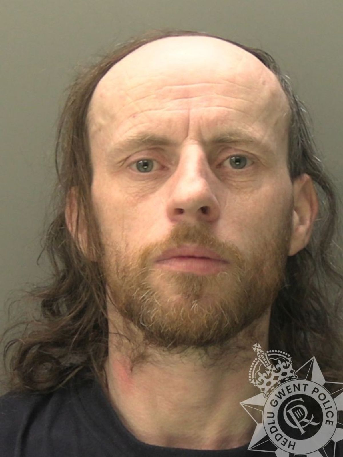 Custody photo of Ashley Coughlan, 42, who is bald and has long brown hair, has a brown beard and is wearing a black t-shirt.