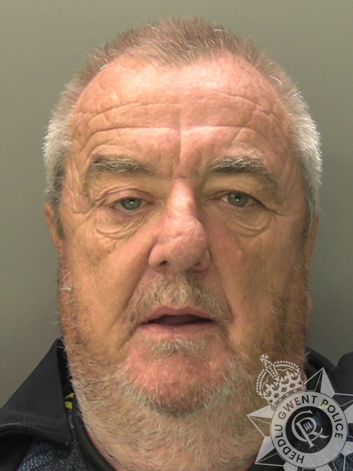 Custody photo of Christopher Preston, 68, who has short grey hair, grey stubble and is wearing a black coat.