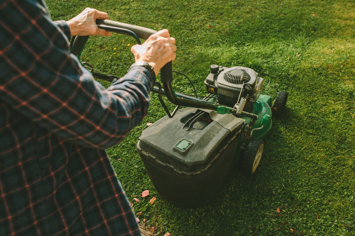A senior woman operates a push lawn mower in a rustic backyard, surrounded by a mix of green grass, landscaped features, and natural elements. Wearing wellington boots and a plaid shirt, she exemplifies active living and a hands-on approach to gardening and home maintenance. Perfect for themes of gardening, senior lifestyle, and rural living.