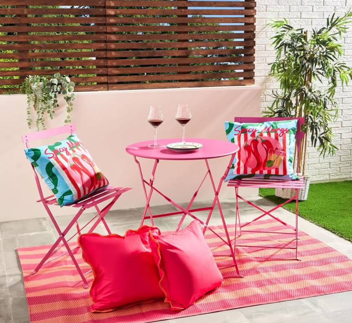 An outdoor patio setting featuring a round pink table accompanied by two vibrantly colored chairs. The table has a white plate placed upon it, and the chairs are adorned with colorful decorative pillows. The patio is bordered by a white wall with wooden blinds and a lush green plant.