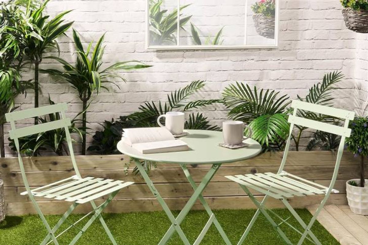 An image depicting an outdoor seating arrangement featuring a round table and two folding chairs. The setting is on a green grassy area, with a white wall and lush greenery in the background. The table is adorned with two white cups, a book, and a cloth, suggesting a peaceful and inviting outdoor dining or relaxation space.