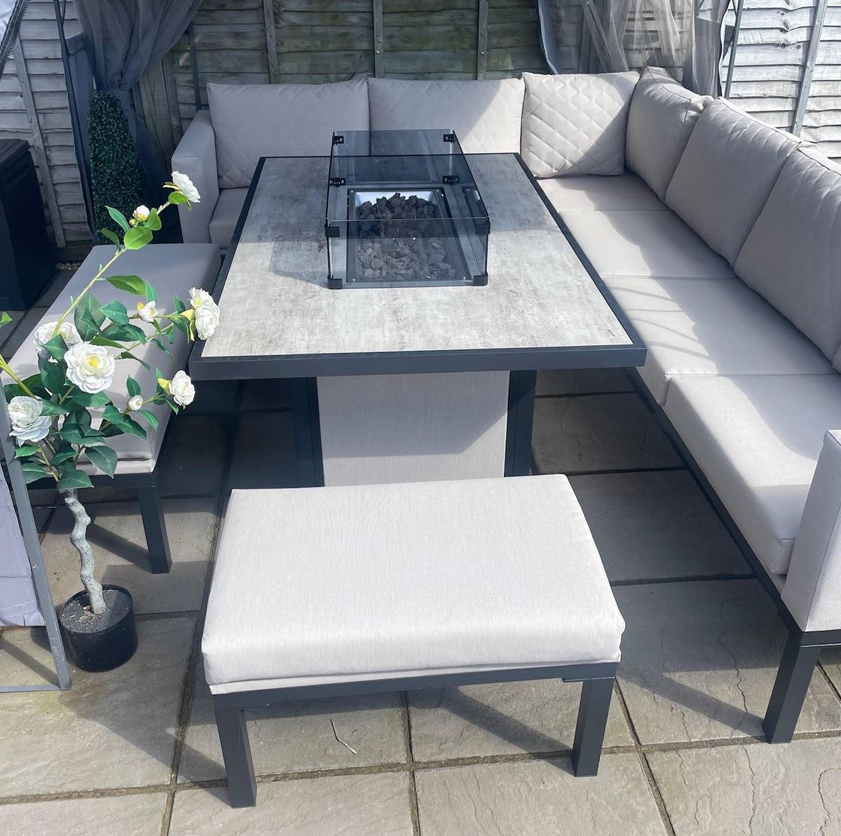 An elegant outdoor seating arrangement featuring a rectangular glass-topped table surrounded by a pair of L-shaped cushioned couches and two cushioned ottomans, adorned with a white floral arrangement on the left side.