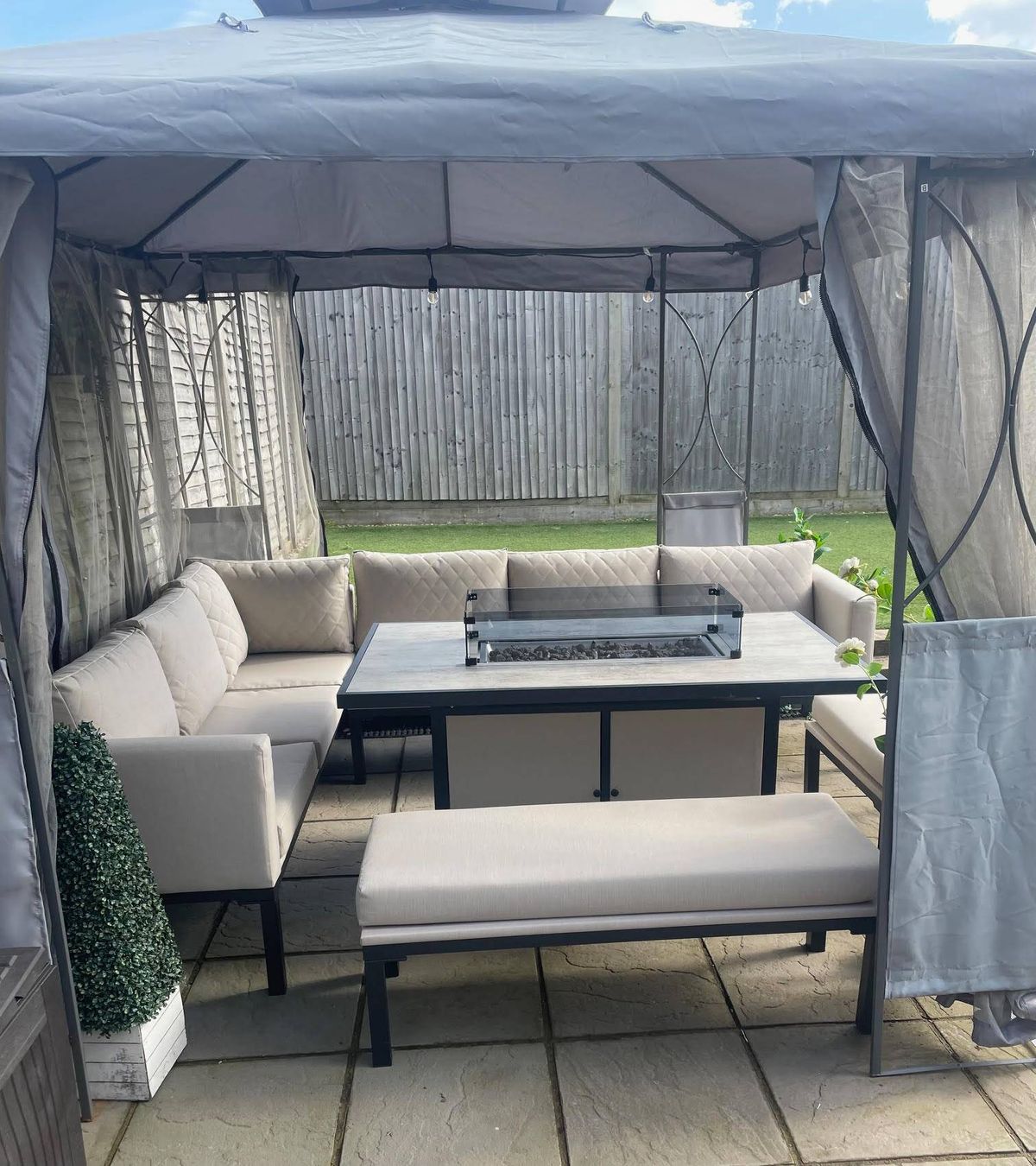 An outdoor seating arrangement featuring a set of white sofas and a central table with a metallic top, surrounded by a paved floor and a potted plant. The area is enclosed by transparent fabric, providing a sheltered environment.