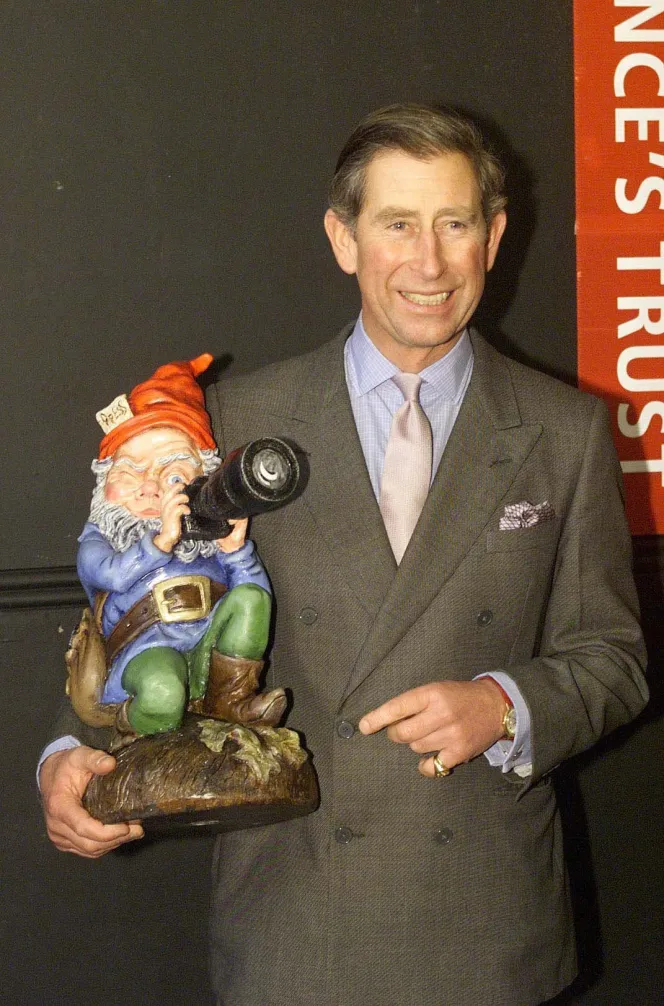 Prince Charles holding a garden gnome wearing a red hat with "Press" written on it and holding a camera.