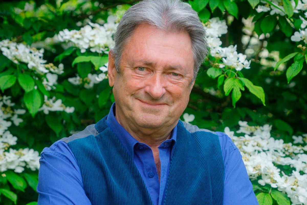 An individual with gray hair is standing and smiling in front of a backdrop of lush greenery and white blossoms. The individual is dressed in a blue long-sleeved shirt and a dark blue vest, exuding a sense of casual elegance.