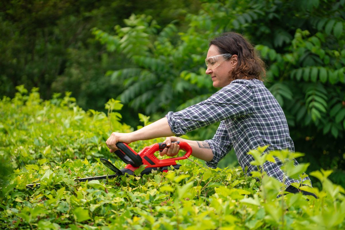A woman wearing protective eyewear and a plaid shirt maintains garden shrubs using a power tool. The serene outdoor setting features lush greenery, trees, and a focus on gardening activities.