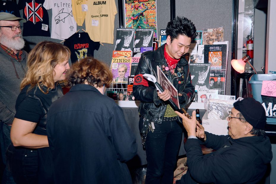 A group of people interact at a merchandise table with punk band shirts, zines, records, and posters displayed in the background.