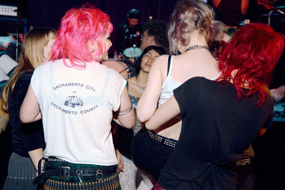 A group of people with colorful hair and punk-style clothing stand together at a music concert, facing the stage.