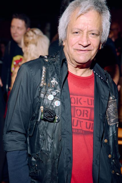 An older man with gray hair wearing a black vest with pins and a red graphic t-shirt, standing indoors at a social event.