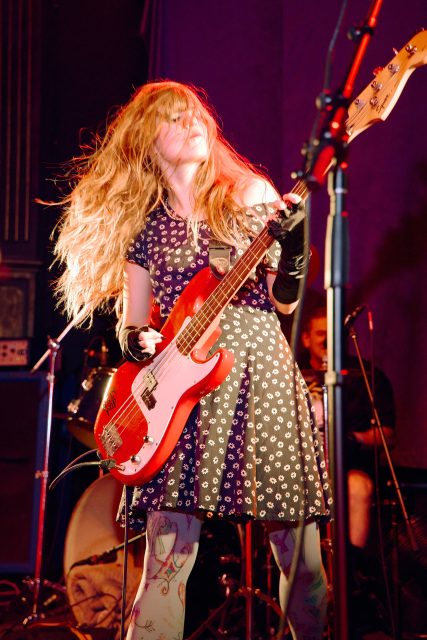 A woman with long hair plays an electric bass guitar on stage, wearing a patterned dress and patterned tights, with stage lights illuminating her.