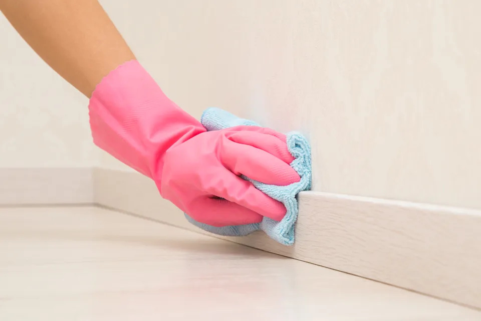 Young adult woman hand in pink rubber protective glove using blue dry rag and wiping light wooden baseboard surface in room at home.