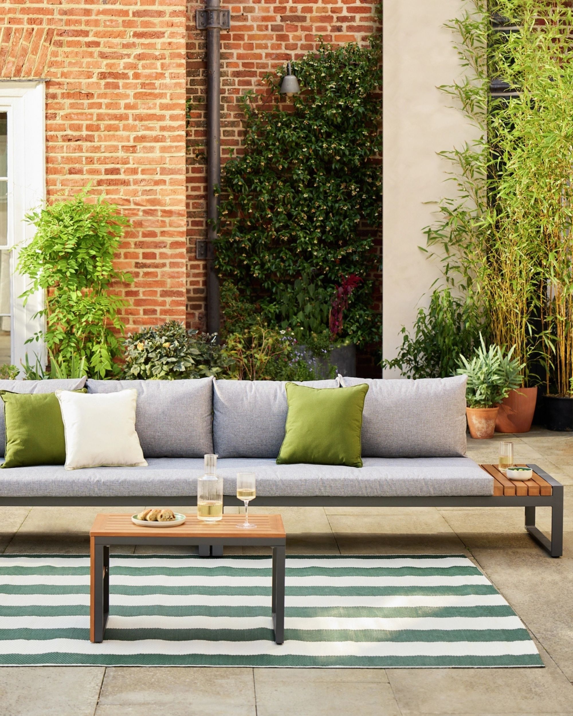 Image of a gray modular outdoor seating seat with a table in front of it that has a wine glass and a carafe. There is a brick house in the background with lots of green plants in front of it.
