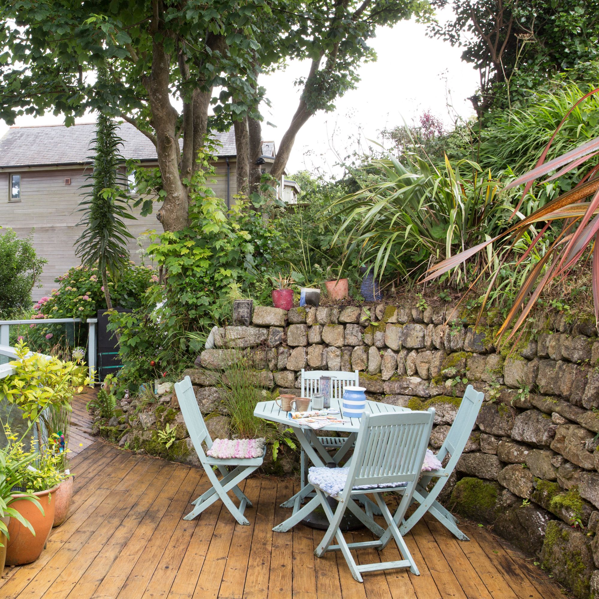 Blue painted wooden garden furniture next to stone wall on wooden decking area with curved edges in garden