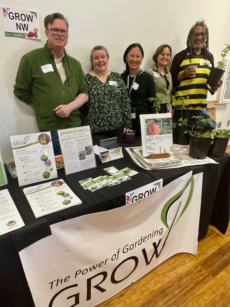 Five people including one person wearing a bee costume, standing in a line smiling behind a table with a banner that says "The Power of Gardening, GROW"