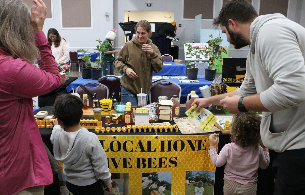 A Table with honey-themed items stand in the middle of a room with more vendors around it. A girl stands, laughing behind the table while parents and children looking at the honey items.