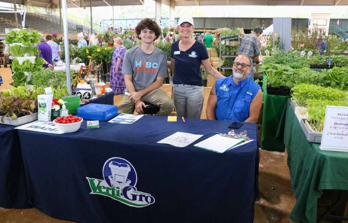 Three people stand behind a table with a tablecloth that reads, "Verti-Gro." Plants surround them in the background.