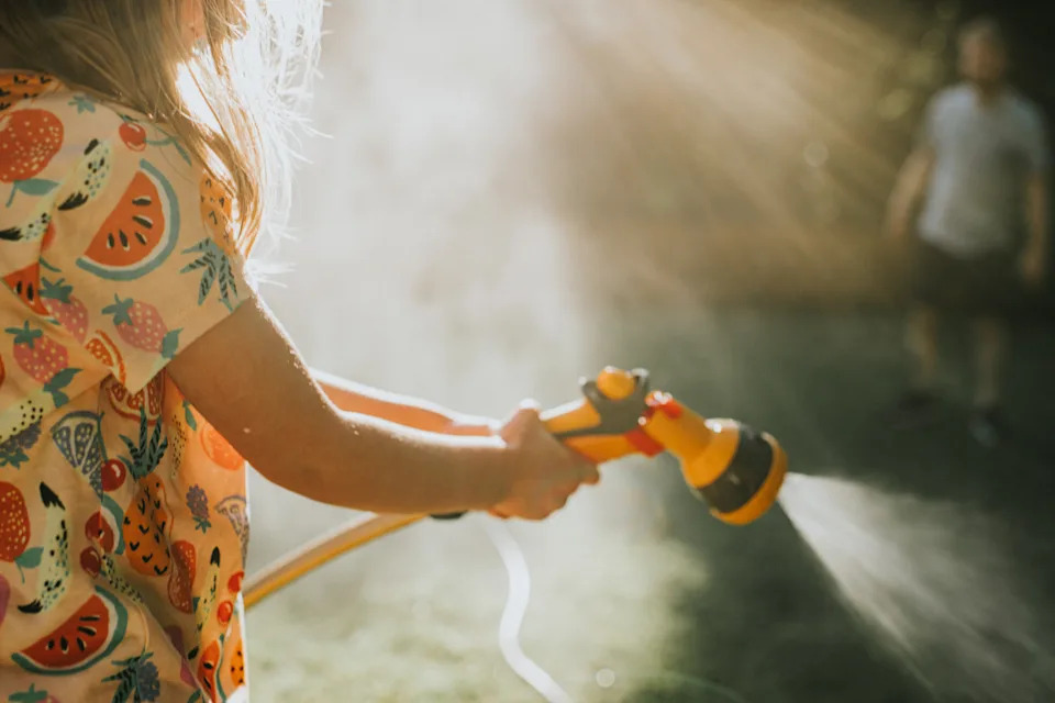 A mischievous girl holds a garden hose with an attachment. She sprays the hose with an attachment.