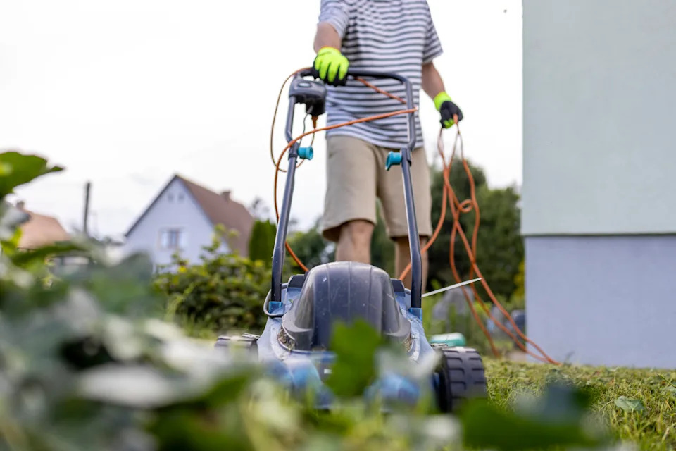 Unrecognisable man in casual clothes and protective gloves mows grass in a garden with a lawnmower.