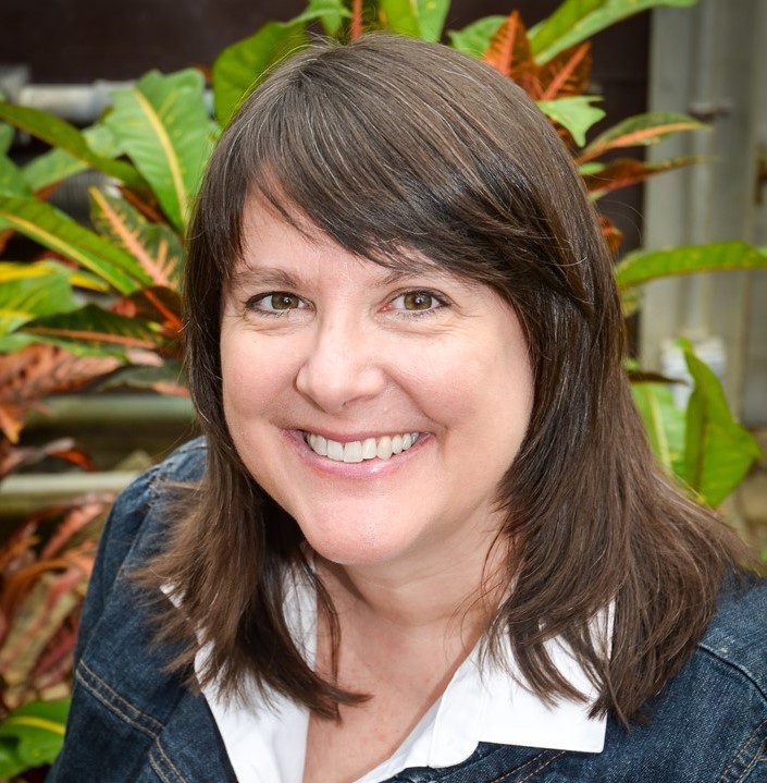 Headshot of woman with brown hair, smiling, in front of plants