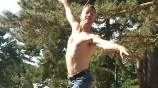 Barechested Karl Stevens, jumps into a dancing pose in a garden