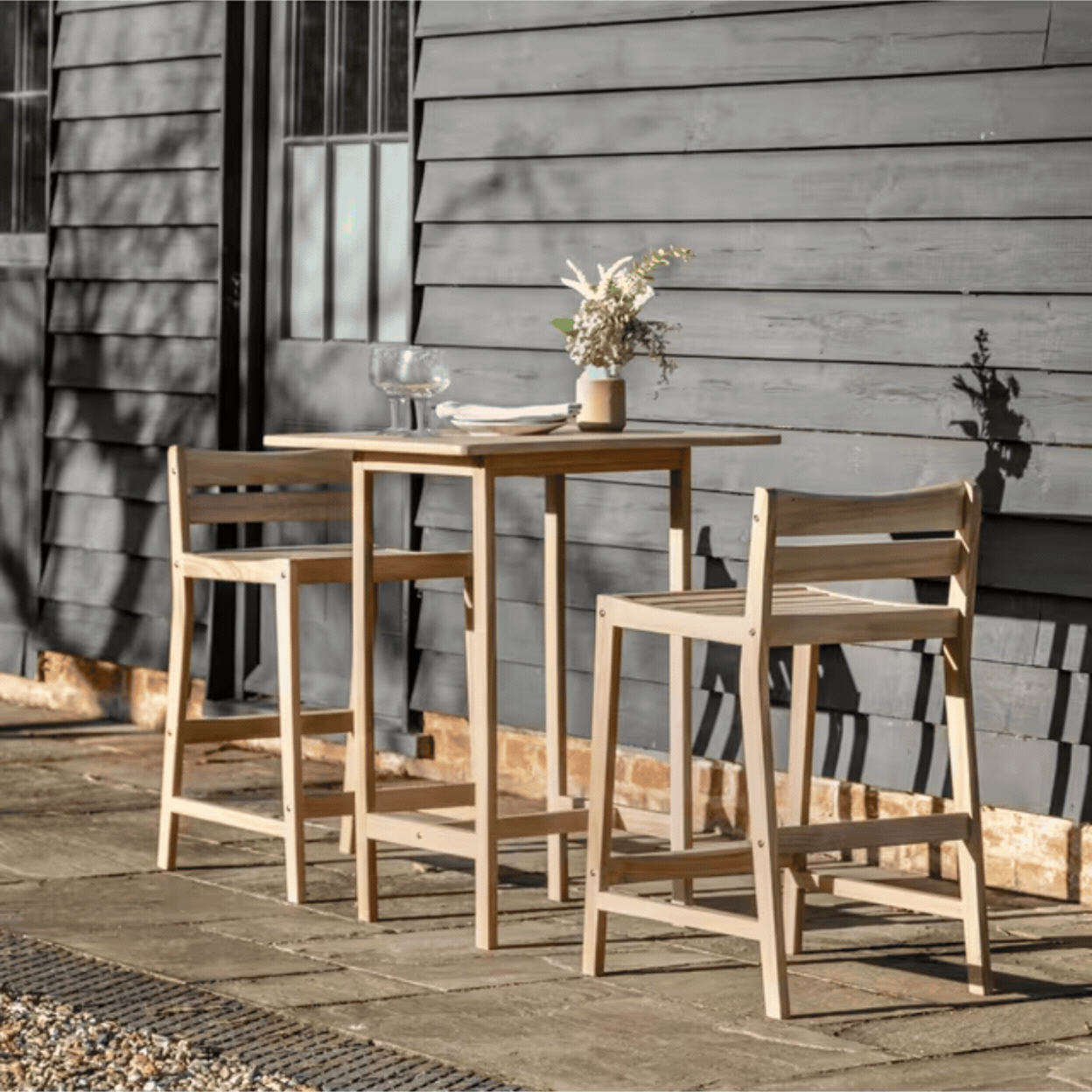 Astakos Acacia Wood 2-Seater Square High Bar Table &amp; Chairs Set, Natural in a rotating gif showcasing it out in the sunshine and as a product against a pale green background.