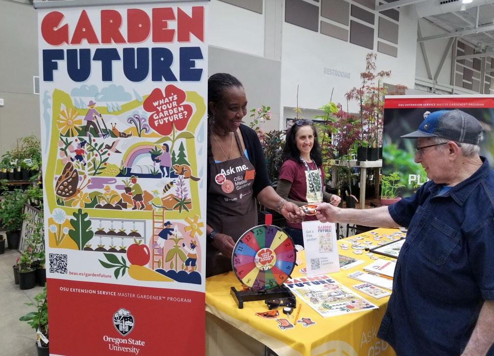 Garden Future helps Oregon gardeners plan for a changing climate Master Gardener volunteer staffs a “Garden Future” booth, handing materials to a visitor beside a colorful display and information table.