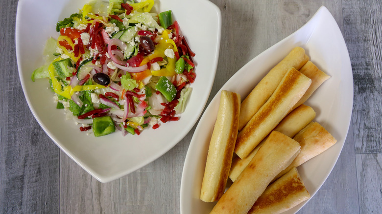 A bowl of breadsticks rests next to a salad on a wooden surface