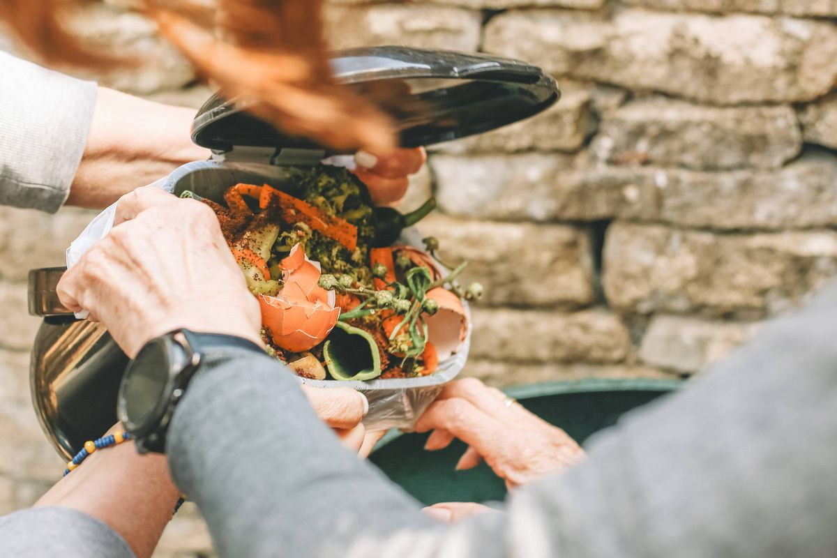 Hands holding kitchen compost container filled with vegetable peelings and eggshells before emptying into outdoor compost bin.